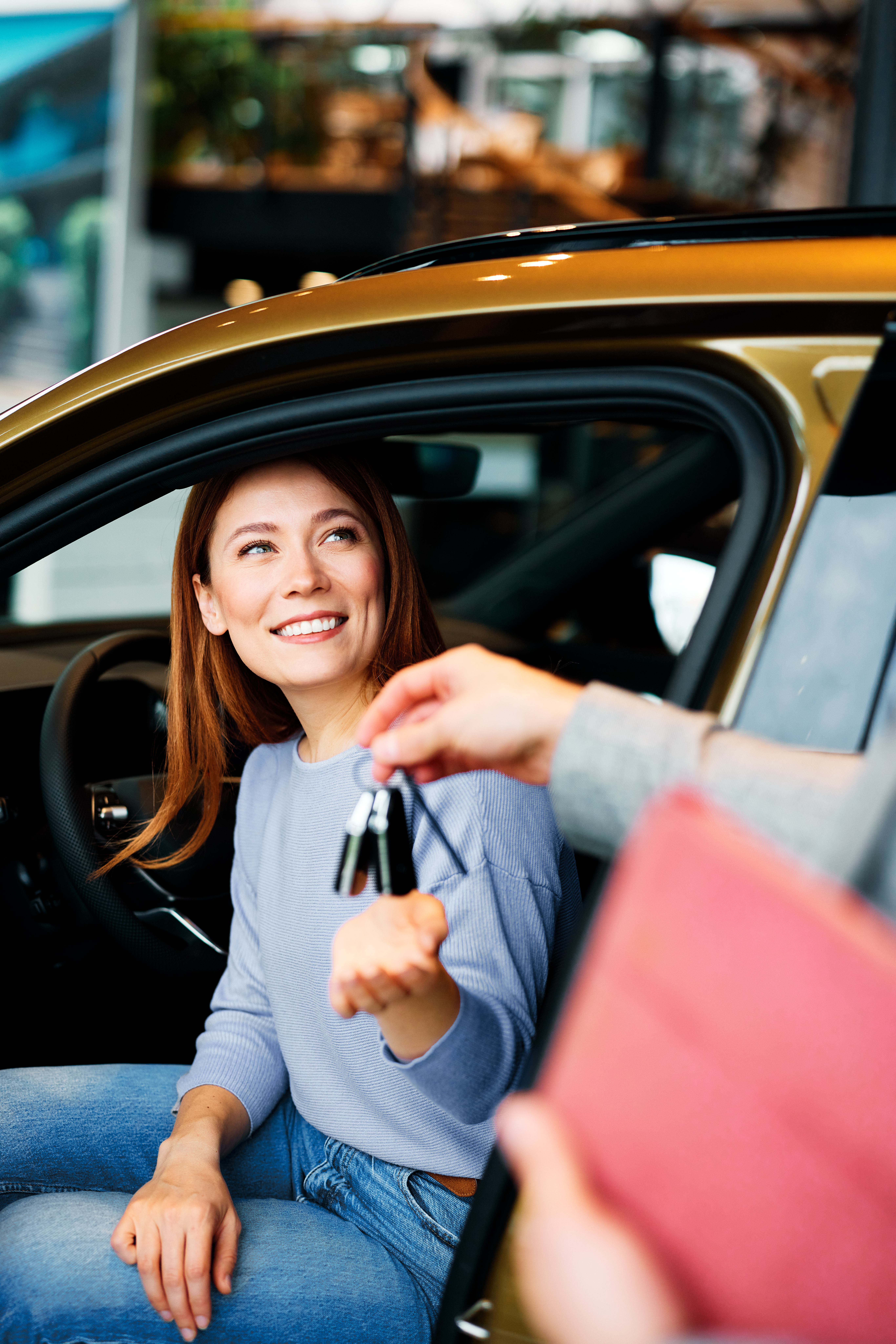 Happy young woman sitting in a new car and receiving keys from a salesman during vehicle purchase at modern car dealership
